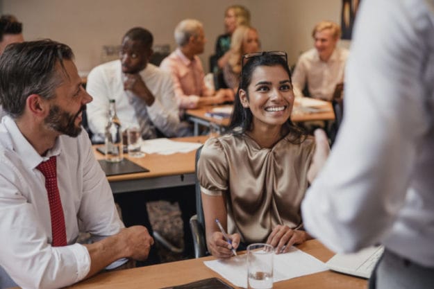 Group of business people sit at desks and face speaker in small conference room