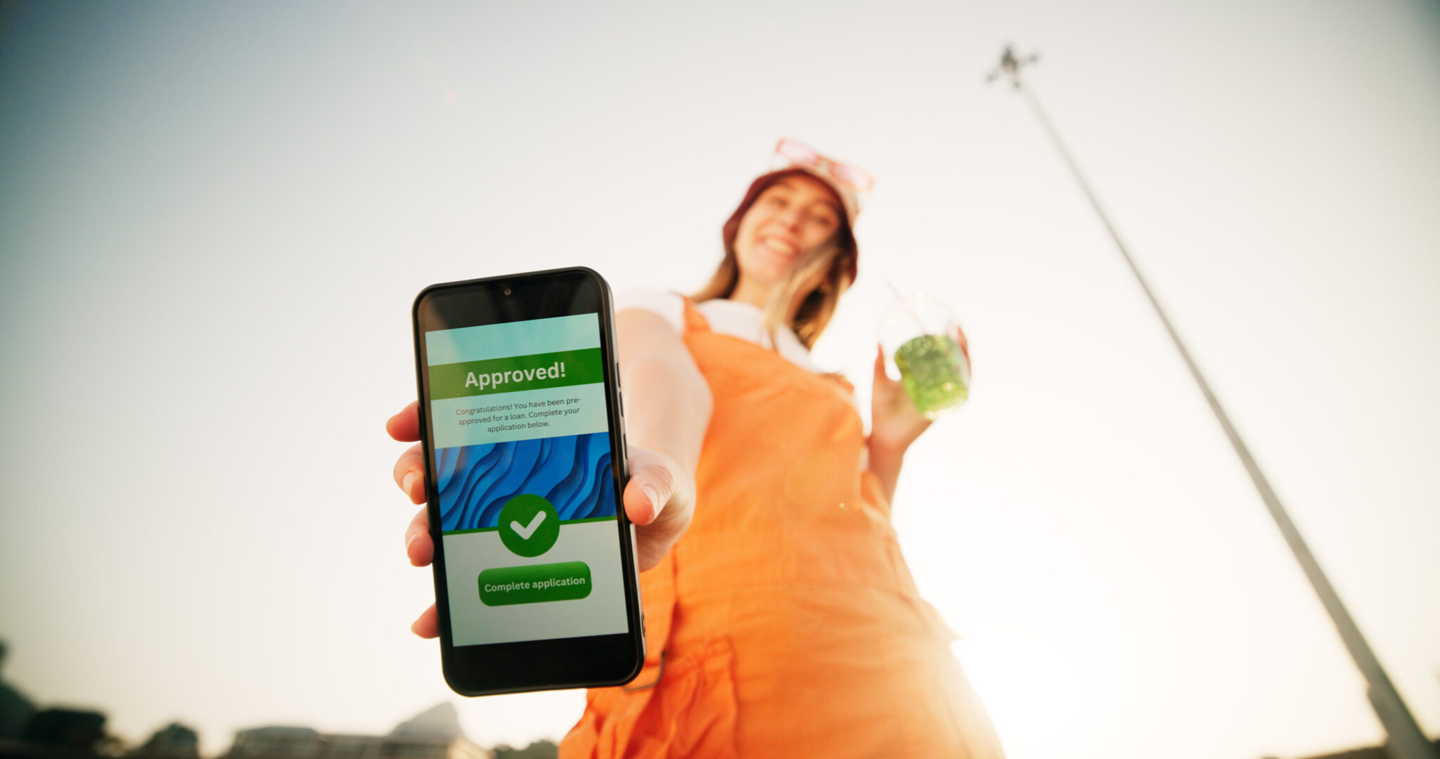 Young woman in the street, holding her phone with a loan approval, with a drink in her hand