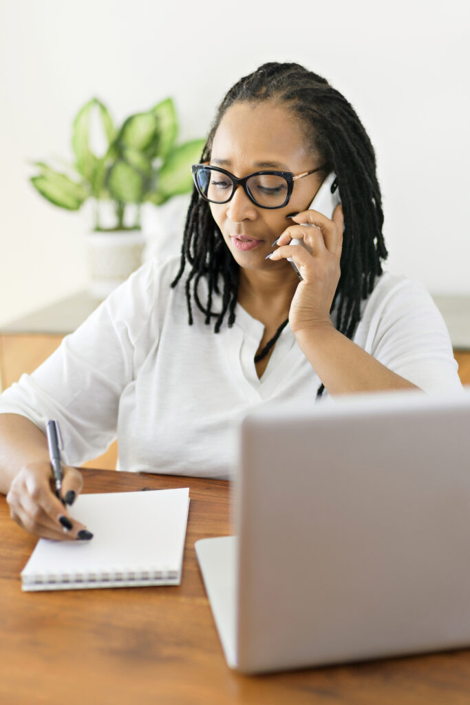 A mature woman using computer in modern kitchen interior, on her phone while taking notes.