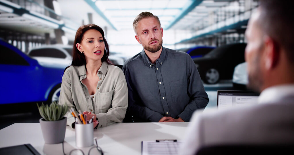 A couple looking stoic while sitting at the car dealership counter, talking with salesperson.