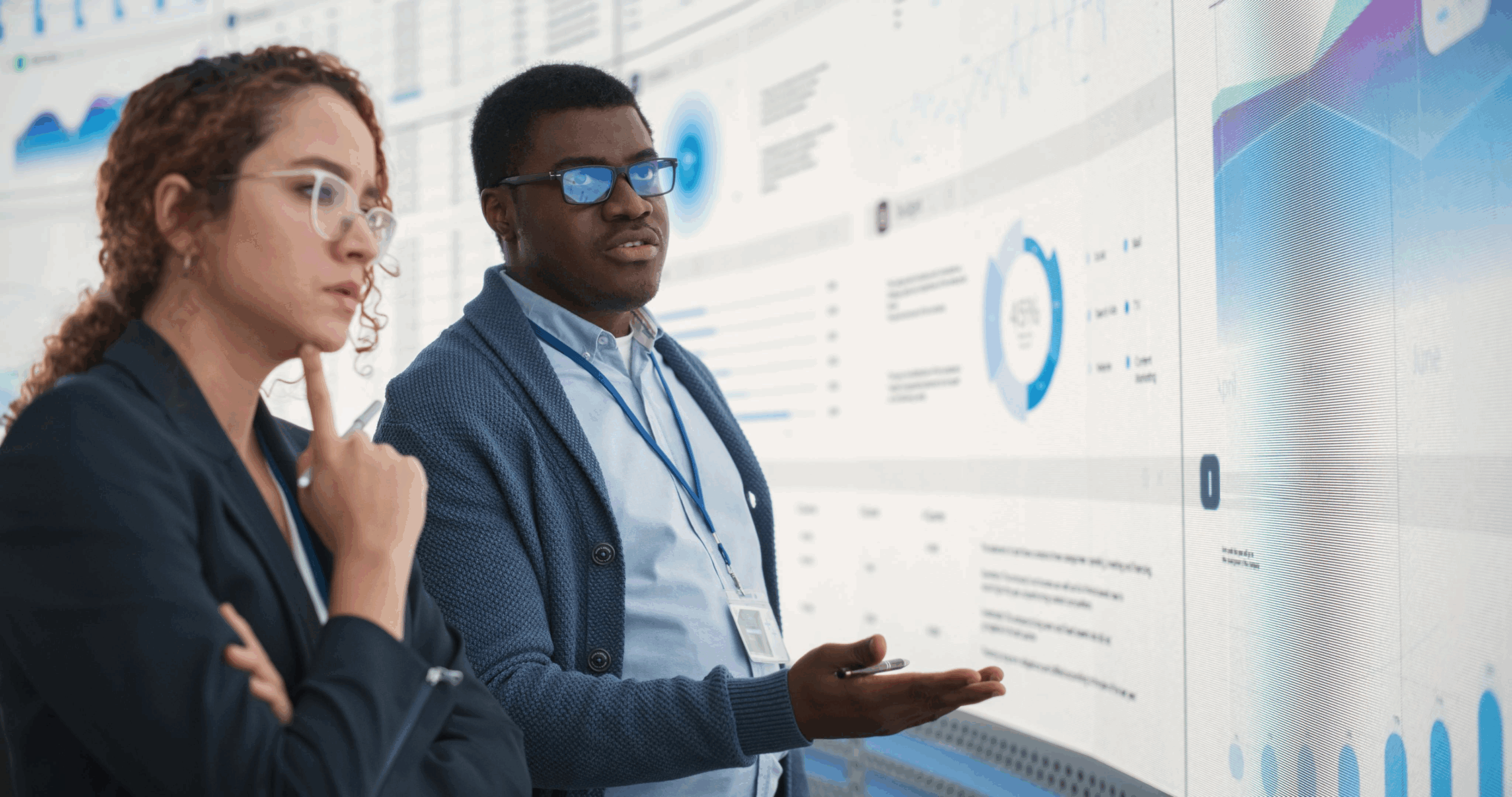 Black Man And Hispanic Woman Working On Business Strategy and Standing Infront of Big Digital Screen With Data. Male And Female Colleagues Discussing Plan For Profit Margins Enlargement In The Office.